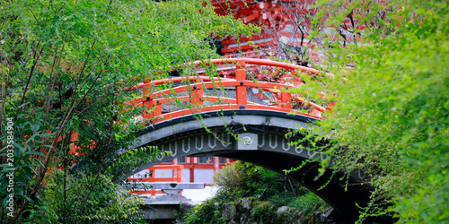 Red arched bridge with orange railing surrounded by green trees at Shinto shrine, Kyoto, Japan