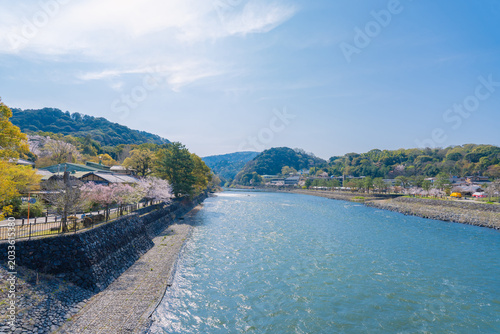Beautiful scenic view of Uji River flowing through Uji city in Kyoto, Japan, during spring season with blooming cherry blossom trees, surrounded by green mountains under a bright clear sky.