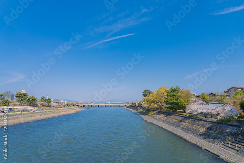 Beautiful scenic view of Uji River flowing through Uji city in Kyoto, Japan, during spring season with blooming cherry blossom trees, surrounded by green mountains under a bright clear sky.
