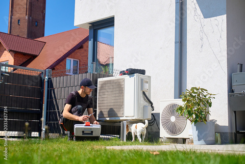 Specialist servicing outdoor heat pump unit while a small dog stands near the equipment. Person inspecting heat pump on lawn near house on sunny day. HVAC service concept