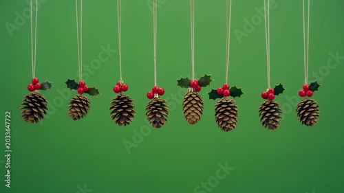 A row of nine pine cone Christmas ornaments with holly and red berries hanging against a green screen chroma key background.