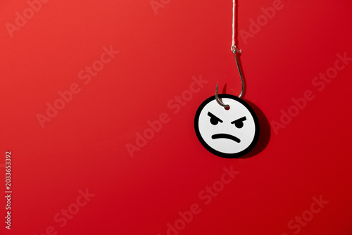 Obraz na płótnie A metallic fishhook piercing an angry face emoji on a red background