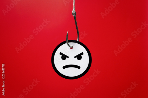 Photography A metallic fishhook piercing an angry face emoji on a red background