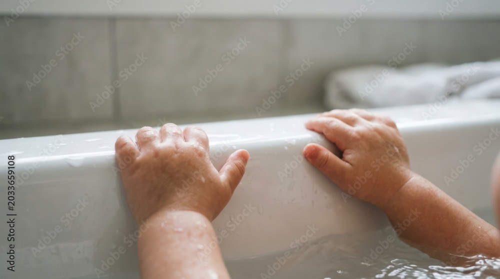 Fototapeta premium Child's hands playing in bathtub with water splashes, enjoying warm bath time experience. Bathtub scene captures joyful moment with little hands gripping edge amidst bubbles and water reflections.