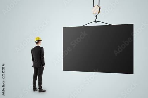 Businessperson in suit and safety helmet looking at a large suspended blank b...