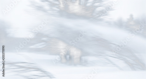 Motion blur view of a snow blower throwing a stream of white snow across a cold, overcast winter landscape with bare trees in the background