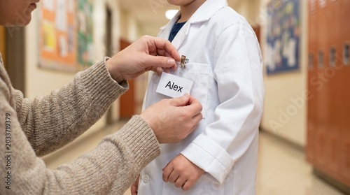 Adult hands attach a name tag to a child wearing a white scientist coat. This scene represents career day or educational roleplay. Ideal for educational materials or parenting blogs.