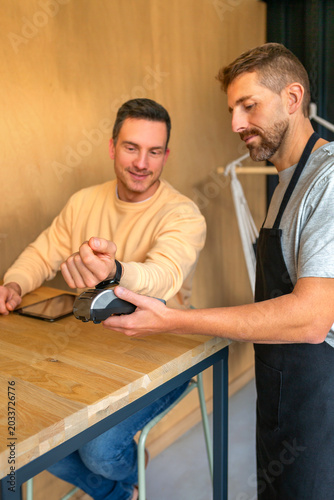 Smiling man with digital tablet taps smartwatch for contactless payment on a terminal held by a bartender at a cafe, showcasing quick, secure tap and go convenience in daily life