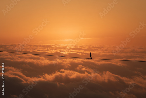 An aerial view of a person walking on a tightrope or scrambling above a vast sea of ​​clouds amidst a sunset.