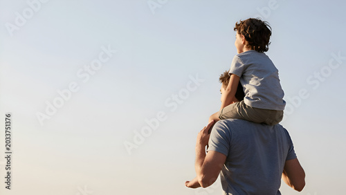 Happy father carrying his little son on shoulders against clear blue sky