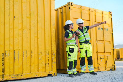 Construction worker in safety helmet and reflective vest standing near yellow container and pointing outdoors in industrial yard