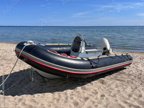 Black inflatable motor boat parked on a sandy beach near calm coastal water under a blue sky