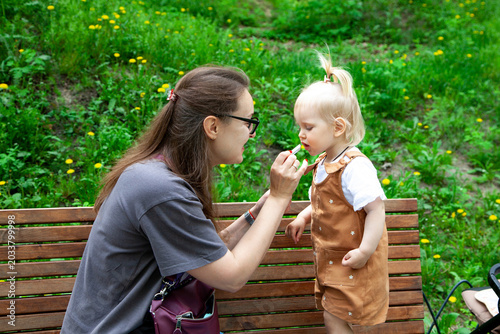 Young mother applying moisturizing lip balm to her little daughter during a walk in a summer park. Caring family moment outdoors, child skincare, motherhood