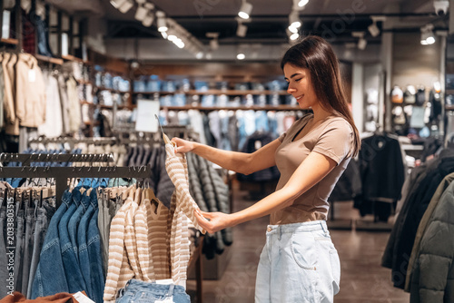 Female shopper examining clothing items in a retail store, surrounded by various garments displayed on racks and shelves in a modern shopping environment