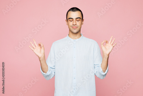 Obraz na plátne Young spiritual middle eastern man wear casual blue cotton shirt hold hands in yoga om aum gesture relax meditate try calm down isolated on plain pastel light pink wall background