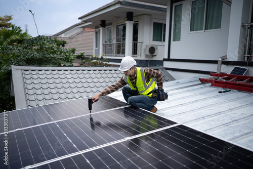 A focused male engineer in safety vest and hardhat installs solar panels with Power Drill on a house roof, ideal for renewable energy and sustainability concepts