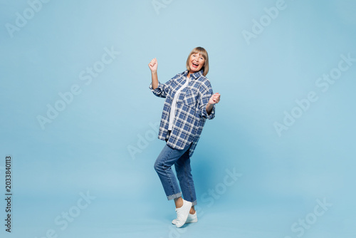 Full body elderly blonde woman 50s years old wear checkered shirt casual clothes doing winner gesture celebrate clench fists say yes raise up leg isolated on plain blue background. Lifestyle concept.