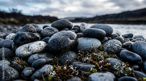 Pebble Shoreline Serenity: A close-up view of smooth, weathered pebbles and moss forms a textured foreground against the backdrop of a calm river and a moody, overcast sky.