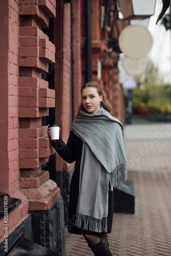 A beautiful young stylish girl in a black coat and a gray scarf walks through the city streets with coffee in her hands