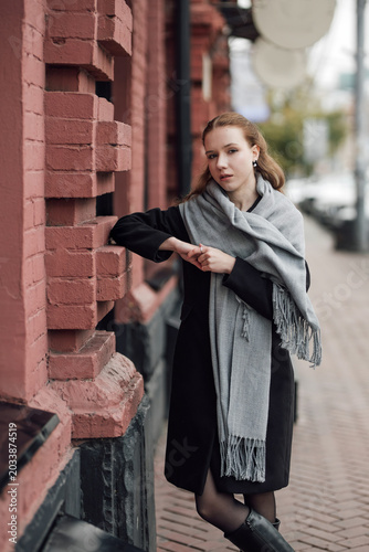 A beautiful young stylish and fashionable girl in a black coat and a gray scarf walks through the city streets