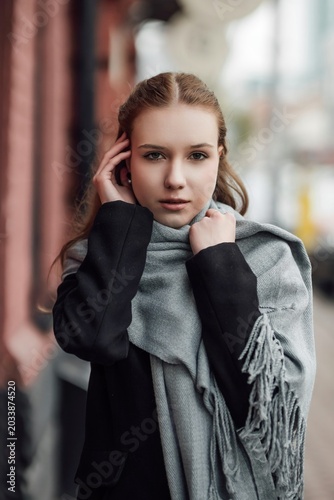 A beautiful young stylish and fashionable girl in a black coat and a gray scarf walks through the city streets