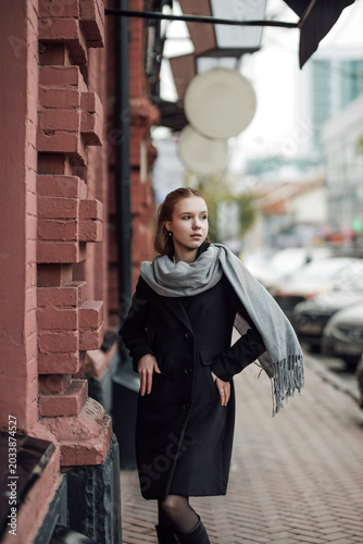 A beautiful young stylish and fashionable girl in a black coat and a gray scarf walks through the city streets