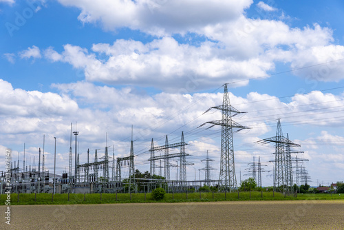 View of Kraftwerk Irsching power plant substation and high-voltage transmission towers standing in a field under a blue sky with clouds in Vohburg an der Donau, Bayern, Germany.