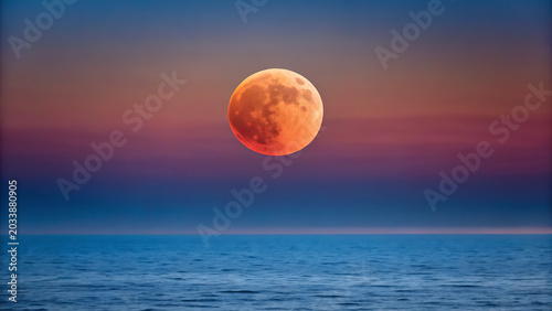 Large orange full moon rising above the calm dark blue ocean water during a twilight sky with soft purple and orange horizon gradients