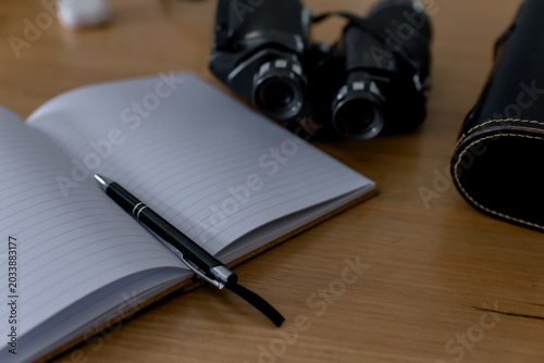Open notebook with pen and binoculars on wooden desk, representing travel planning, observation, research, adventure preparation, journaling, exploration, and outdoor discovery concept.