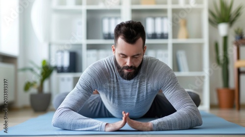 A bearded man performs a seated yoga stretch on a mat in a bright home office eyes closed in concent n embodying work from home wellness and mental clarity