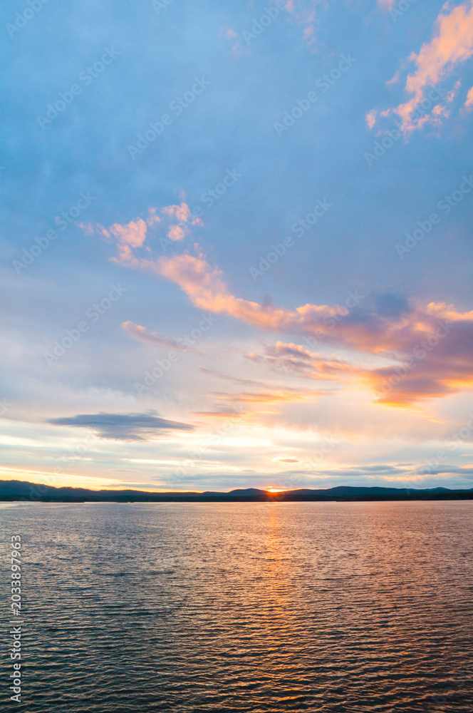 Fototapeta premium Picturesque summer sunset water landscape, dramatic sky over the rippled water surface and the sun setting behing the mountains