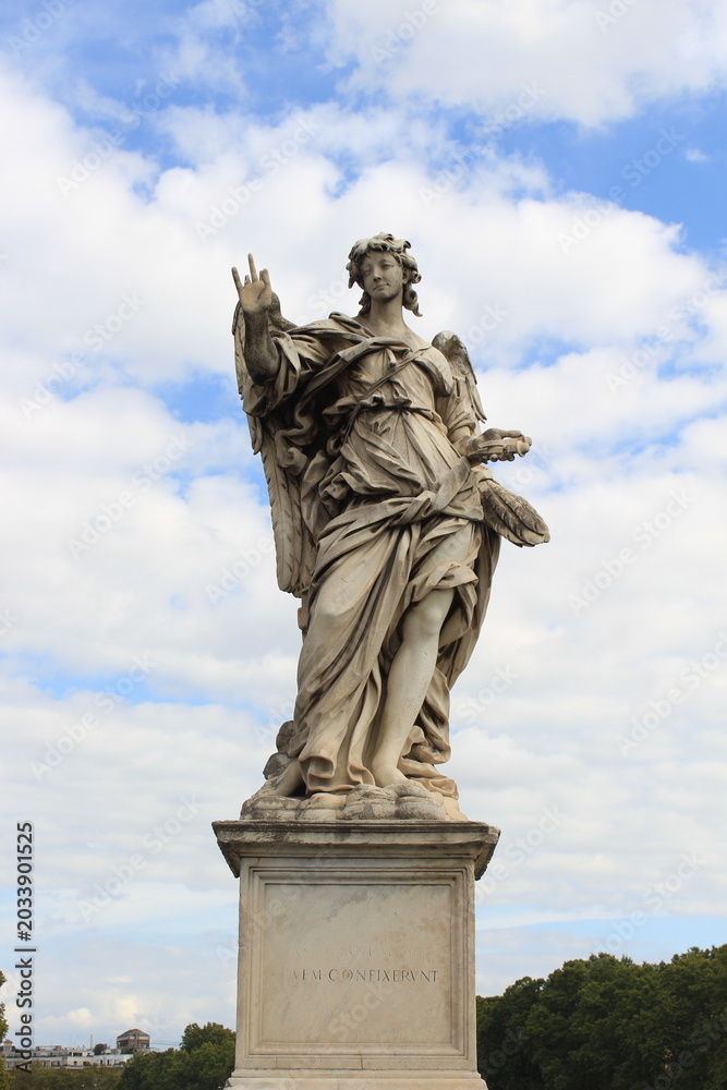Fototapeta premium A detailed marble statue of an angel with flowing drapery set against a bright, partly cloudy sky, embodying classical baroque art.