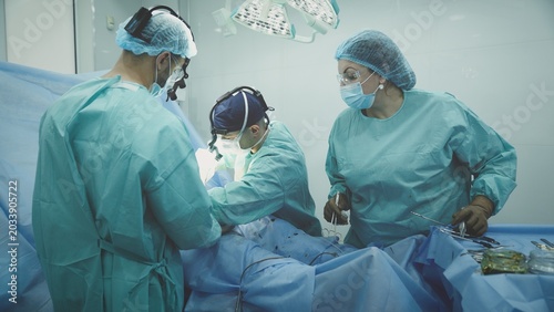 Three clinic workers in protective uniform surgeons doctors operating patient medicine health care procedure. Nurse woman help men practitioners operate at table malignant tumor remove surgery health