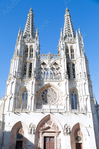 A detailed view of the towers of Burgos Cathedral, showcasing its Gothic spires, ornate stone carvings, and pointed arches against a bright blue sky.