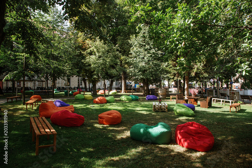 View of colorful bean bags and wooden benches scattered on a green lawn under the shade of lush trees in a public park Sremska Mitrovica, Vojvodina, Serbia.