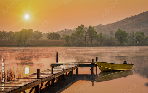Fotografía Peaceful sunrise over Petpenoun Lake in Cameroon, Africa, featuring a wooden dock and small boat reflected on calm water