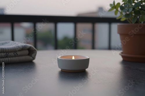 Candle holder made of white ceramic material цшер  a small burning flame sits on a dark table surface. Table on an outdoor balcony holds a candle, a folded blanket, and a potted plant.