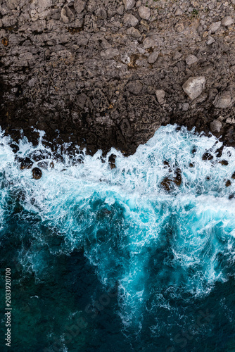 A dramatic top-down aerial view of turquoise ocean waves crashing against a rugged rocky coastline. 