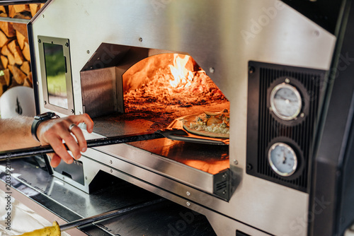 Cook slides a prepared pizza on a peel into a hot stainless oven with visible flames, firewood, and temperature gauges