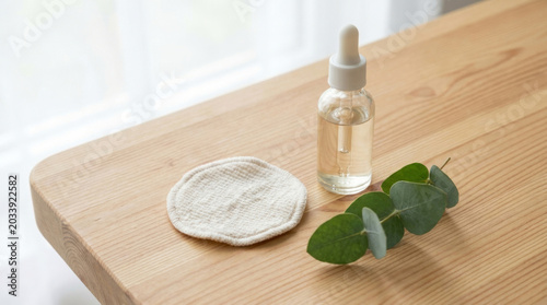 A serene beauty setup featuring a glass dropper bottle of oil, a cotton pad, and fresh eucalyptus leaves on a wooden table.