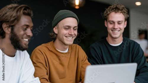 Three smiling young men collaborate around a laptop in a casual indoor workspace with shallow focus.