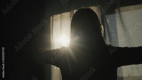 Woman silhouette slowly opens curtains revealing bright morning sun, illuminating room and offering hope and new beginning.