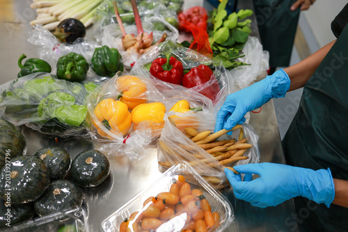 Focused diligent worker wearing blue glove sorting fresh vegetable into plastic packaging ensuring food safety and hygiene during preparation on metal table