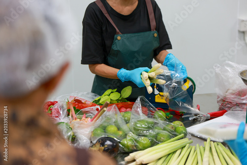 Focused worker wearing green apron and blue glove carefully packing fresh vegetable like lemongrass and lime into plastic bag for commercial kitchen food preparation and local market distribution