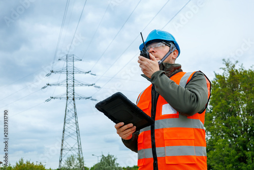 Worker using a portable radio for real-time reporting and damage prevention during a pre-storm site survey. Utility company technician or inspector evaluating electrical grid infrastructure. 