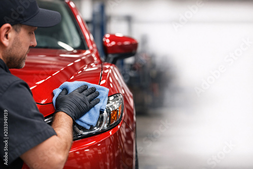 A mechanic wearing gloves and a black shirt washes a red car in an auto body shop.