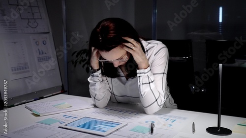 Slika na platnu Businesswoman clutching her head in a dim office, surrounded by paperwork and a