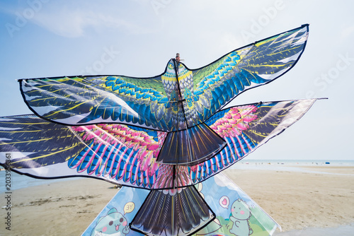 Soaring Duo: Two intricately designed bird-shaped kites, one with vibrant blue and green feathers and the other with pink and purple hues, sit on a sandy beach under a bright blue sky.