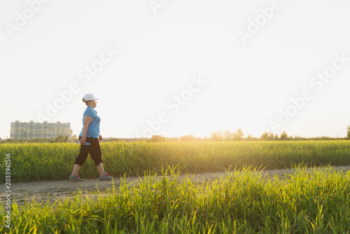 Active senior woman in sport clothes hiking on outdoor in the field with bottle of water. Summer season. Selective focus. Wellness, healthy lifestyle