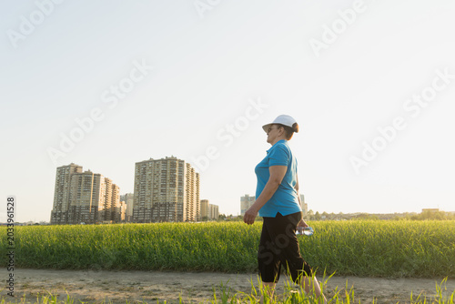 Active senior woman in sport clothes hiking on outdoor in the field with bottle of water. Summer season. Selective focus. Wellness, healthy lifestyle